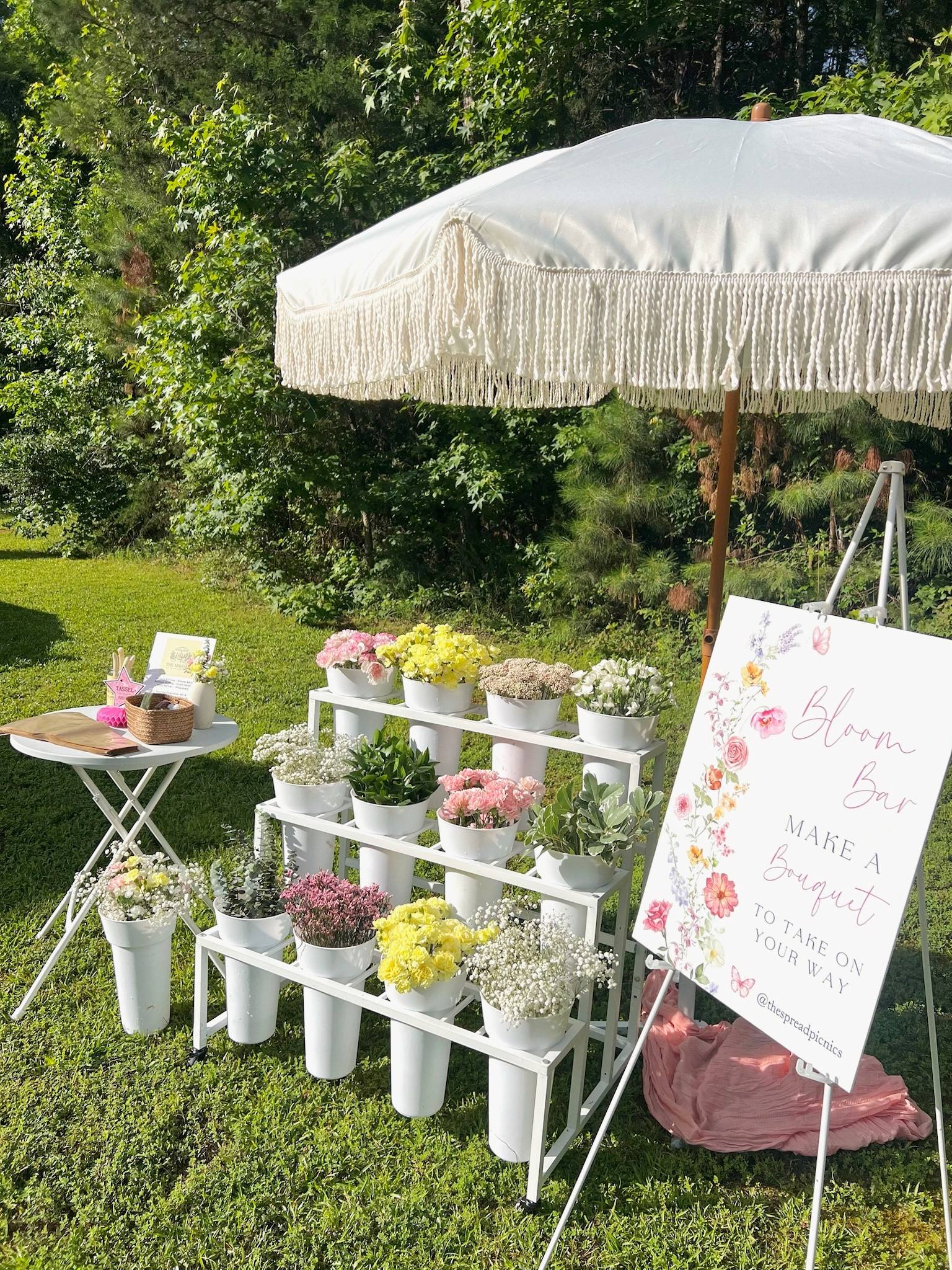 A white tiered Bloom Bar stand of flower buckets under a cream fringed umbrella, beside a wrap table and a Make a Bouquet sign on an easel, on a lawn by pine trees