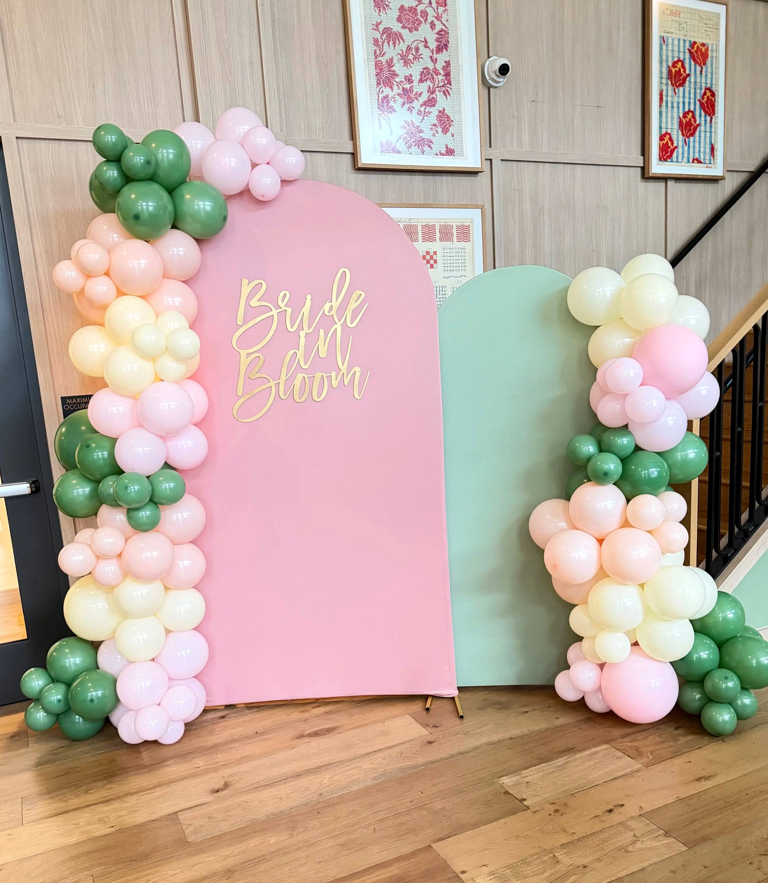 A pink arch backdrop lettered Bride in Bloom beside a sage arch, framed by pink, cream and green balloon garlands